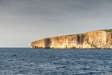 Rugged limestone cliffs of Comino Island in Malta overlooking the deep blue waters of the Mediterranean Sea under a clear afternoon sky. Travel to Malta.