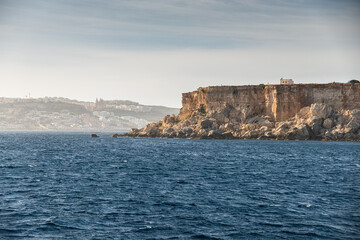Rugged limestone cliffs of Comino Island in Malta overlooking the deep blue waters of the Mediterranean Sea under a clear afternoon sky. Travel to Malta.