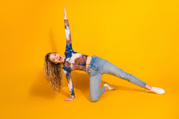 Smiling young woman with blonde hair performs a dynamic stretch on a bright yellow background wearing casual denim and tie dye top for a playful fashion fitness moment