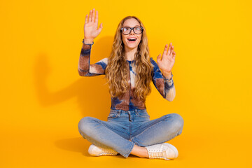Young woman with glasses sits cross legged in yellow studio posing joyfully