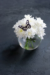 glass vase with bouquet of white chrysanthemum flowers and decorative butterfly on dark background. symbol of tenderness, sadness, revival life, mourning. copy space. template for design