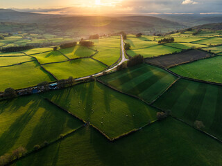 Aerial view of sunbeams streak across the patchwork quilt of vibrant green fields intersected by a solitary road, Brecon, Wales, United Kingdom.