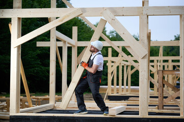Construction worker installing wooden beam at house frame construction site. Carpenter in safety gear building timber structure for new home outdoors.