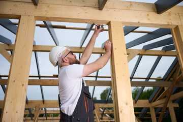 Construction worker measuring wooden beam with tape measure at building frame. Builder in safety helmet working on wooden house construction site outdoors.