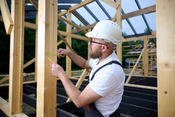 Construction worker measuring wooden beam with tape measure at building frame. Builder in safety helmet working on wooden house construction site outdoors.