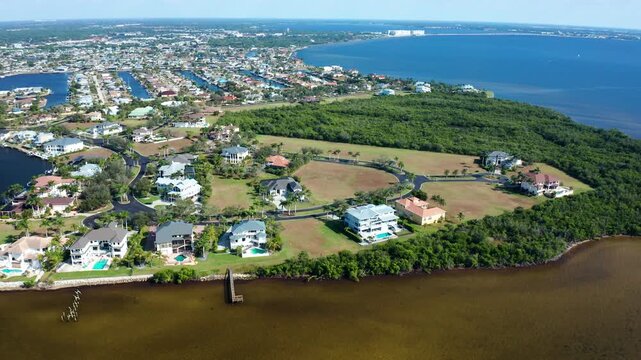Waterfront luxury homes with swimming pools and green lawns are situated on a peninsula surrounded by bay waters and dense foliage, with a distant cityscape visible in Florida.