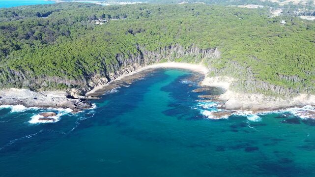 Drone aerial landscape of sandy beach cove with ocean waves crashing along rocky reef and headland covered by bushland forest national park reserve at Malua Bay South Coast Australia travel nature