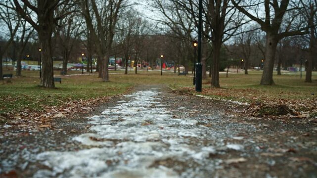Icy park pathway with wet pavement, patchy ice and fallen leaves in early winter, calm urban seasonal transition background. Lateral movement.
