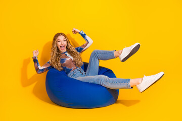 Young girl in jeans on a blue beanbag smiles excitedly in bright yellow studio background