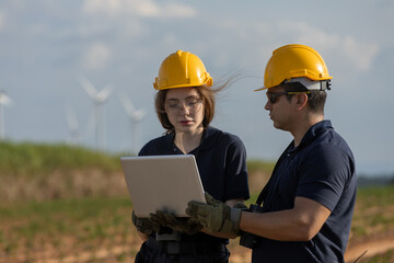 Two engineer wearing yellow helmets analyzing data on laptop in wind farm, symbolizing digital monitoring, renewable energy management, teamwork and sustainable technology in the field.