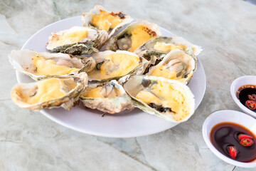 A white plate contains baked oysters with melted yellow cheese topping next to two small bowls of dark sauce with sliced red chili on a marbled table.