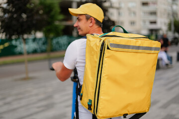 A person operates a scooter and carries a large yellow bag on their back as they move through a city setting with buildings around them