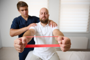 Physiotherapist guiding male patient with resistance band exercise for shoulder rehabilitation in clinic. Physical therapy session focused on injury recovery, strengthening, mobility improvement