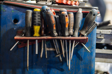 Close-up of many used screwdrivers with handles of different colors and textures hanging on a red shelf or holder in a workshop or garage.