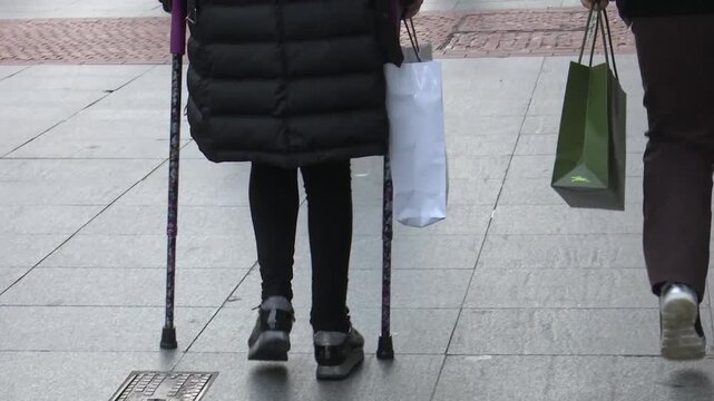 Piernas de personas caminando por la calle con bolsas de compras y una persona con muleta en temporada de rebajas.