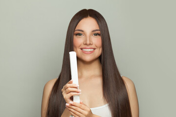 Glorious woman brunette with long straight smooth hairstyle using hair straightener iron on light grey background