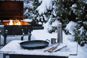 Burning fire in outdoor barbecue grill with frying pan, camp thermos and utensils on snowy table in...