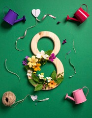 Background for Women's Day, a wooden "8", from which flowers and plants grow, around the 8 lie ribbons and strings and watering cans.