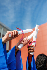 Diverse students proudly raising their rolled diplomas with pink ribbons towards a blue sky