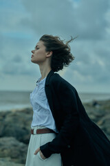 Windy coastal scene featuring a woman in a dark coat over a white shirt, belt visible, standing on...