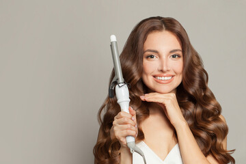 Posing woman with long wavy hairstyle using hair curling tongs iron, indoor portrait.