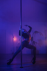 Athletic woman performing pole dance moves in studio with dramatic colored lighting