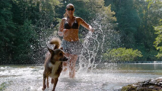 SLOW MOTION, PORTRAIT: Woman in a bikini top and shorts runs through a shallow river, making water splashes. Two dogs are running alongside her, fully immersed in joyful and energetic outdoor activity