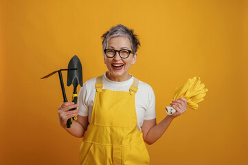 Senior woman gardener in bright yellow overalls smiles while holding gardening tools and gloves...