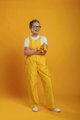 Senior woman wearing yellow overalls and glasses stands confidently against a vibrant yellow background, showcasing a cheerful and positive attitude in a playful setting