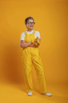 Senior woman wearing yellow overalls and glasses stands confidently against a vibrant yellow background, showcasing a cheerful and positive attitude in a playful setting
