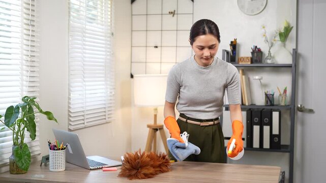 An Asian woman wearing orange rubber gloves wipes a wooden desk with a microfiber cloth and spray bottle in a modern home office, focusing on cleanliness, hygiene, and workspace maintenance.