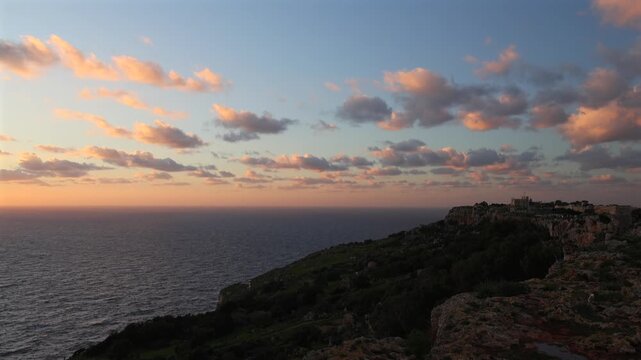 Golden hour sunset at the rugged Dingli Cliffs in Malta, with warm light illuminating the rocky terrain and small clouds over the Mediterranean Sea. 4k video
