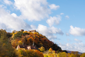 Autumn landscape of a historic castle and church on a hill surrounded by colorful fall foliage. Beautiful sunny day with golden trees and a blue sky with soft white clouds