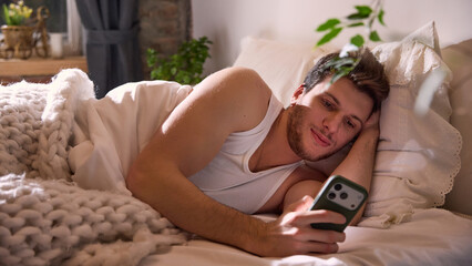 Man lying on bed using smartphone in cozy bedroom. Concept of mobile technology at home, digital comfort, app based services, online communication, everyday tech lifestyle.