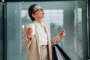 Businesswoman in stylish beige suit holds coffee cup and shopping bags, looking up thoughtfully in...