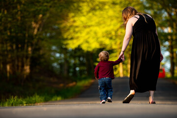Obraz premium A mother and her little boy walk hand in hand on an open road in the sunset. The mom and child seen from behind depict a mother's love and care, perfect for Mother's Day and family content.