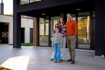 Fototapeta premium Happy family, including a father, mother, and young daughter, smiling and standing together outdoors in front of a contemporary house, symbolizing homeownership, real estate, and togetherness