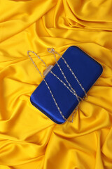 Top view of elegant blue satin clutch bag with silver chain on vibrant yellow silk fabric. Luxury evening purse on draped golden textile. High-contrast fashion flat lay product photography.