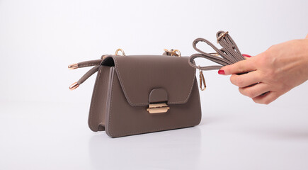 Elegant brown pebble leather crossbody bag with gold hardware. Woman's hand with red nails holding a matching detachable shoulder strap. Professional fashion accessory isolated on white background.