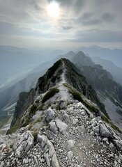 Panoramic view of a mountain ridge hiking trail with a bright sun peeking through clouds, leading through a vast, hazy alpine landscape.