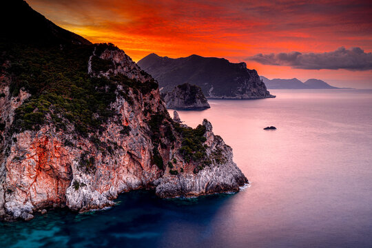 Picturesque red sunrise over the Ionian island Corfu, Gyali beach. Rocky coast with mountains on a holiday island in Greece. The most beautiful viewpoints in Europe.
