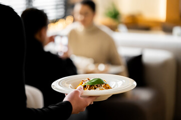 A waiter serving a gourmet pasta dish to customers in a blurred, elegant restaurant setting with warm lighting.