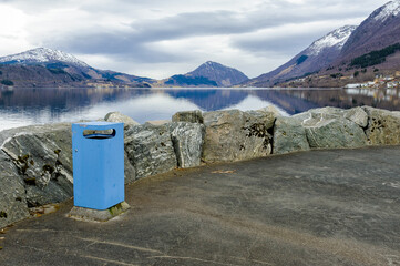 A blue trash bin stands on a rocky shore by the lake. Mountains rise in the distance under a cloudy sky. The water reflects the surrounding landscape.