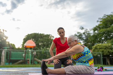 Young female trainer assisting an elderly woman with leg stretching exercises outdoors