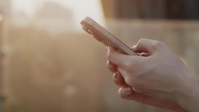 Crop view of young woman hands hold modern smartphone on sunset city street. Female texts to friend by mobile phone tapping touchscreen outdoors. Digital communication technology