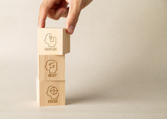 Hand stacking wooden blocks with icons of Unplug, Reset, and Focus. Concept for mental health, digital detox, wellbeing, and work life balance. Great for business productivity and self care.