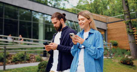 Portrait of man and woman in casual outfits walking outdoors in a city park while using their...