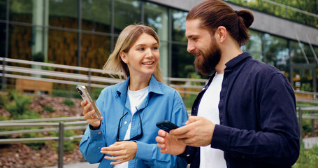 Naklejka premium Two young adults man and woman strolling in a park while checking their mobile phones, sharing something on the screen and smiling during a friendly conversation