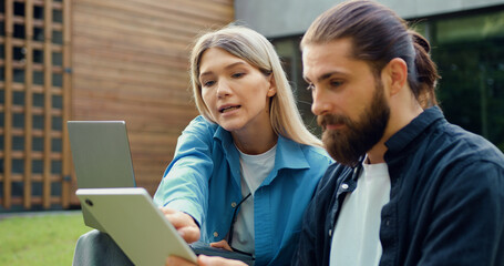 Side view of two colleagues sitting on grass outdoors, the woman pointing at the mans laptop screen while explaining, collaborating in an IT focused work session.