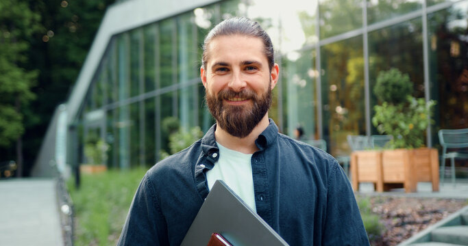 Handsome office worker with laptop, standing in cityscape and giving a friendly smile to the camera.